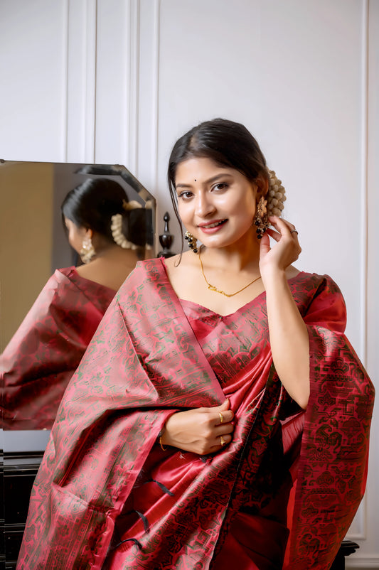 Woman wearing a red silk saree with intricate brocade patterns and a matching blouse, standing gracefully beside a vintage black dresser with a mirror in a softly lit studio featuring white paneled walls and elegant decor.