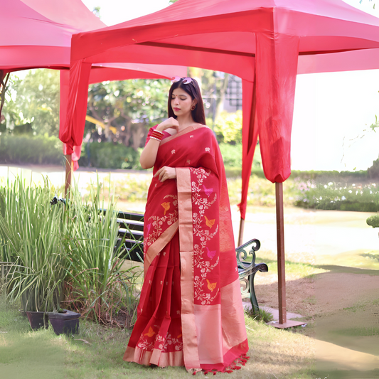 Woman wearing a red saree with peach border and floral embroidery, posing outdoors under a red canopy with green plants and a bench in the background.