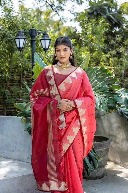 Woman wearing a red handloom silk saree with gold zari border and geometric patterns, paired with a sleeveless blouse and traditional jewelry, standing outdoors on a concrete platform surrounded by lush green plants and garden lighting.