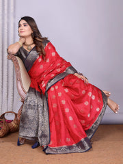 Woman wearing a striking red silk saree with intricate silver and black zari border, detailed buti motifs, and a matching black blouse. She is accessorized with traditional jewelry and bangles, posing elegantly in a contemporary studio setting with woven baskets of rose petals, sheer white curtains, and a neutral gray backdrop.