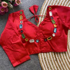 Red blouse embellished with square and round mirror work, featuring multicolored embroidered borders, stylish back tie-up closure, and three-quarter sleeves. Displayed flat on a textured grey surface alongside a woven mat and fresh flowers, highlighting its festive and contemporary ethnic style.