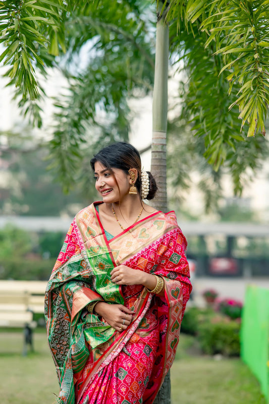 Woman wearing a red saree with intricate zari work and a green blouse, standing outdoors near a tree.