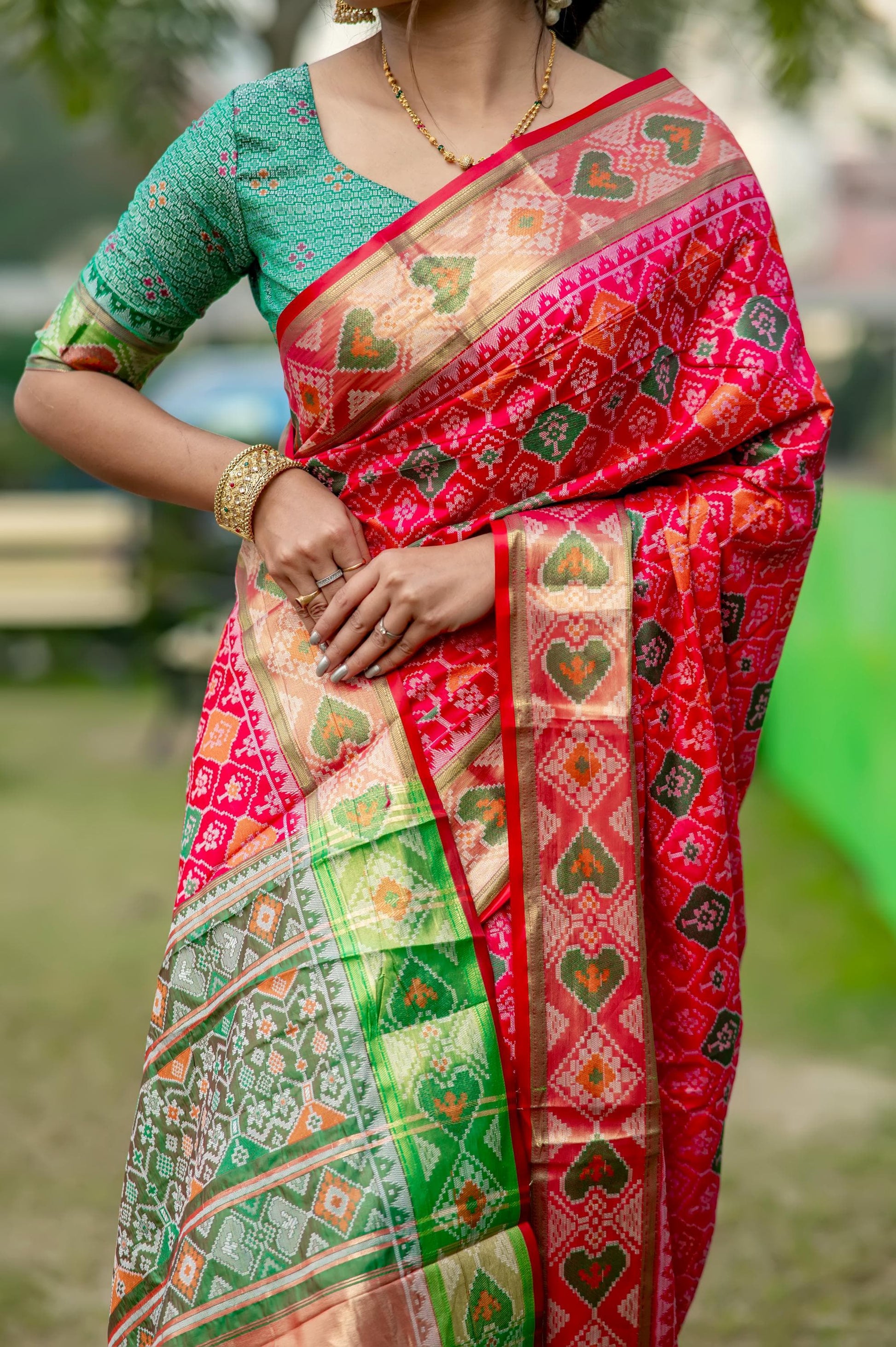 Woman wearing a red blouse and a saree with pink and green floral zari work, standing outdoors with hands clasped.