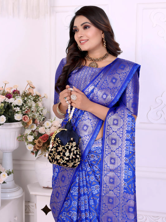 Woman wearing an elegant royal blue Banarasi bandhani saree adorned with intricate silver zari border and traditional tie-dye motifs, paired with a matching blouse and statement jewelry. She poses gracefully in a classic white room decorated with ornate wall panels and vases filled with pastel and rose-toned artificial flowers.