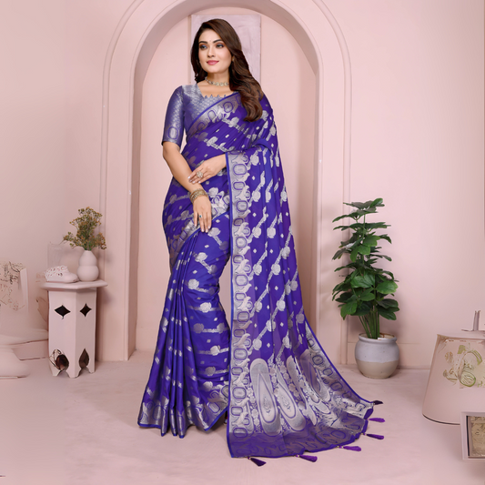 Woman wearing a royal blue Banarasi saree decorated with intricate silver zari motifs and an ornate border with tassel details, paired with a matching textured blue blouse and styled with traditional jewelry and bangles. She poses gracefully against a pastel arched backdrop, featuring minimalist decor including a white pedestal with vases and a potted green plant, highlighting an elegant festive ethnic look.