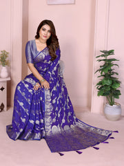 Woman wearing a royal blue Banarasi saree decorated with intricate silver zari motifs and an ornate border with tassel details, paired with a matching textured blue blouse and styled with traditional jewelry and bangles. She poses gracefully against a pastel arched backdrop, featuring minimalist decor including a white pedestal with vases and a potted green plant, highlighting an elegant festive ethnic look.