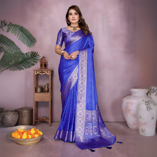 Woman wearing a royal blue Banarasi silk saree with intricate silver zari border and geometric woven motifs, paired with a matching blouse and gold jewelry. She stands gracefully in a contemporary pastel-toned studio, surrounded by minimalist decor including a wooden side table with brass vessels, marigold flowers in a bowl, green foliage, and large white ceramic vases.