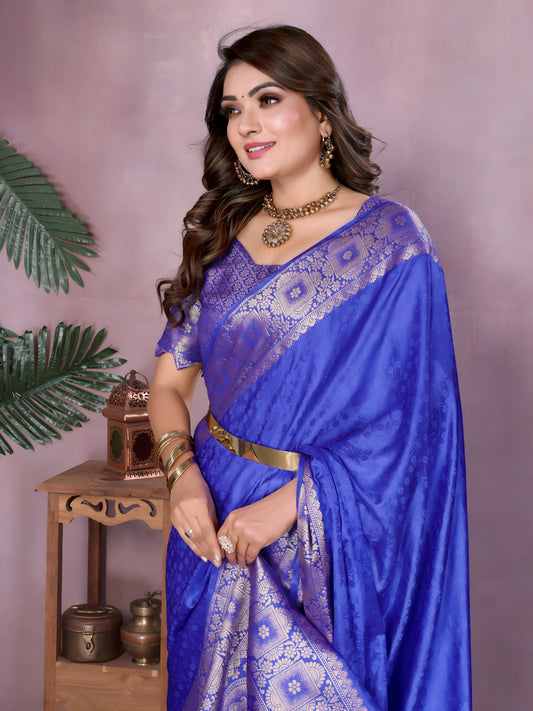 Woman wearing a royal blue Banarasi silk saree with intricate silver zari border and geometric woven motifs, paired with a matching blouse and gold jewelry. She stands gracefully in a contemporary pastel-toned studio, surrounded by minimalist decor including a wooden side table with brass vessels, marigold flowers in a bowl, green foliage, and large white ceramic vases.