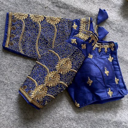 Traditional royal blue silk blouse featuring intricate gold embroidery and beadwork with floral and paisley motifs on the sleeves and neckline, displayed flat on a textured surface with a bunch of artificial flowers and a woven mat in the background.