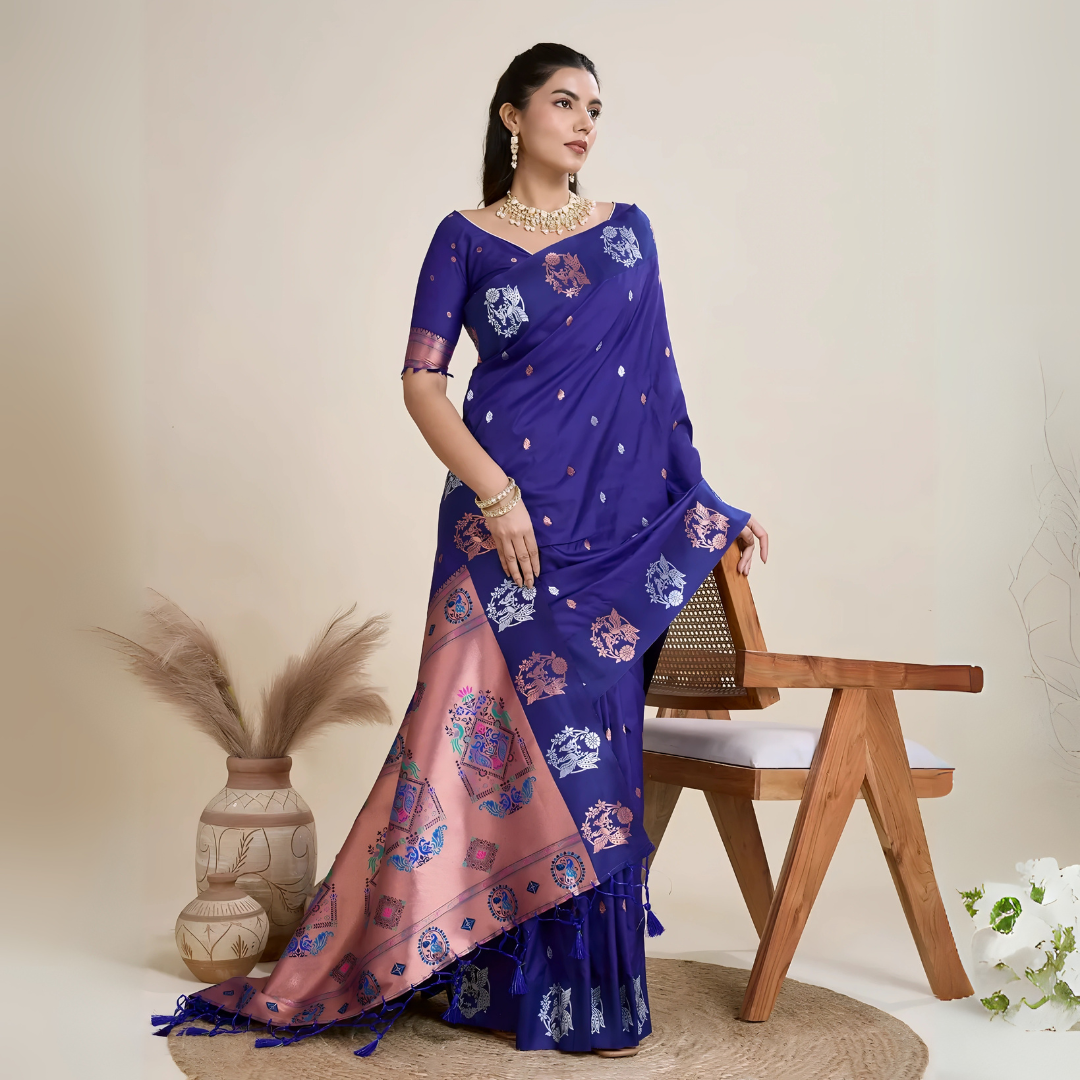Woman wearing a royal blue silk saree adorned with delicate woven motifs and a contrasting pink pallu featuring intricate designs, paired with a matching blouse and traditional jewelry, elegantly posing beside a wooden chair in a minimalist studio setting with neutral decor, ceramic vases, and pampas grass.