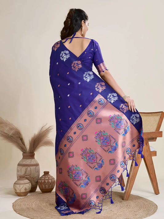 Woman wearing a royal blue silk saree adorned with delicate woven motifs and a contrasting pink pallu featuring intricate designs, paired with a matching blouse and traditional jewelry, elegantly posing beside a wooden chair in a minimalist studio setting with neutral decor, ceramic vases, and pampas grass.