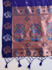 Woman wearing a royal blue silk saree adorned with delicate woven motifs and a contrasting pink pallu featuring intricate designs, paired with a matching blouse and traditional jewelry, elegantly posing beside a wooden chair in a minimalist studio setting with neutral decor, ceramic vases, and pampas grass.