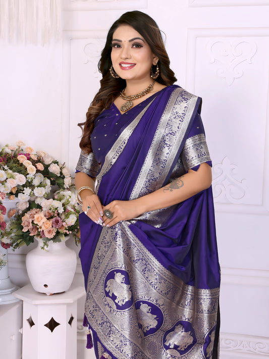 Woman draped in a royal blue silk saree with an elegant silver zari border featuring traditional motifs and tassel accents, styled with a matching blouse and gold jewelry. She stands gracefully in a sophisticated white floral-themed room decorated with classic wall panels and ornate flower arrangements.