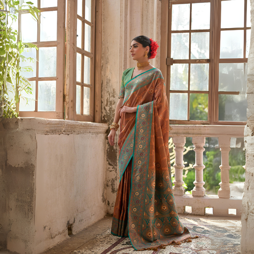 Woman wearing a rust orange silk saree with intricate gold motifs and a turquoise blue border, paired with a green blouse and traditional gold jewelry, standing gracefully in a vintage room with large antique wooden windows and natural light streaming.