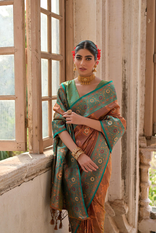 A woman models a striking Bronze Brown Banarasi Silk Saree with all-over Zari weaving. The saree has a wide, ornate Sea Green/Teal border featuring traditional Zari weaving in gold and rust tones. She wears a mint green blouse and gold Kundan jewelry, posing by a large antique window.