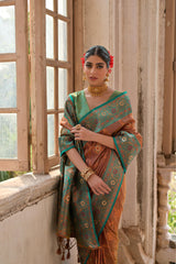 Woman wearing a rust orange silk saree with intricate gold motifs and a turquoise blue border, paired with a green blouse and traditional gold jewelry, standing gracefully in a vintage room with large antique wooden windows and natural light streaming.