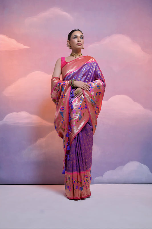 Woman wearing a silver Banarasi silk saree with an intricate pink floral zari border and pallu, paired with a pink blouse and traditional jewelry, posing in a pastel-themed studio with dreamy cloud backgrounds