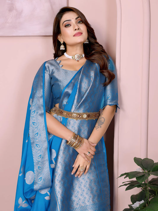 Woman wearing a sky blue silk saree with elegant silver zari border and floral motifs, paired with a matching blouse and traditional silver jewelry. She stands gracefully in front of a pastel arched alcove in a contemporary studio, with a small white table, minimal decor, and a green potted plant completing the setting.