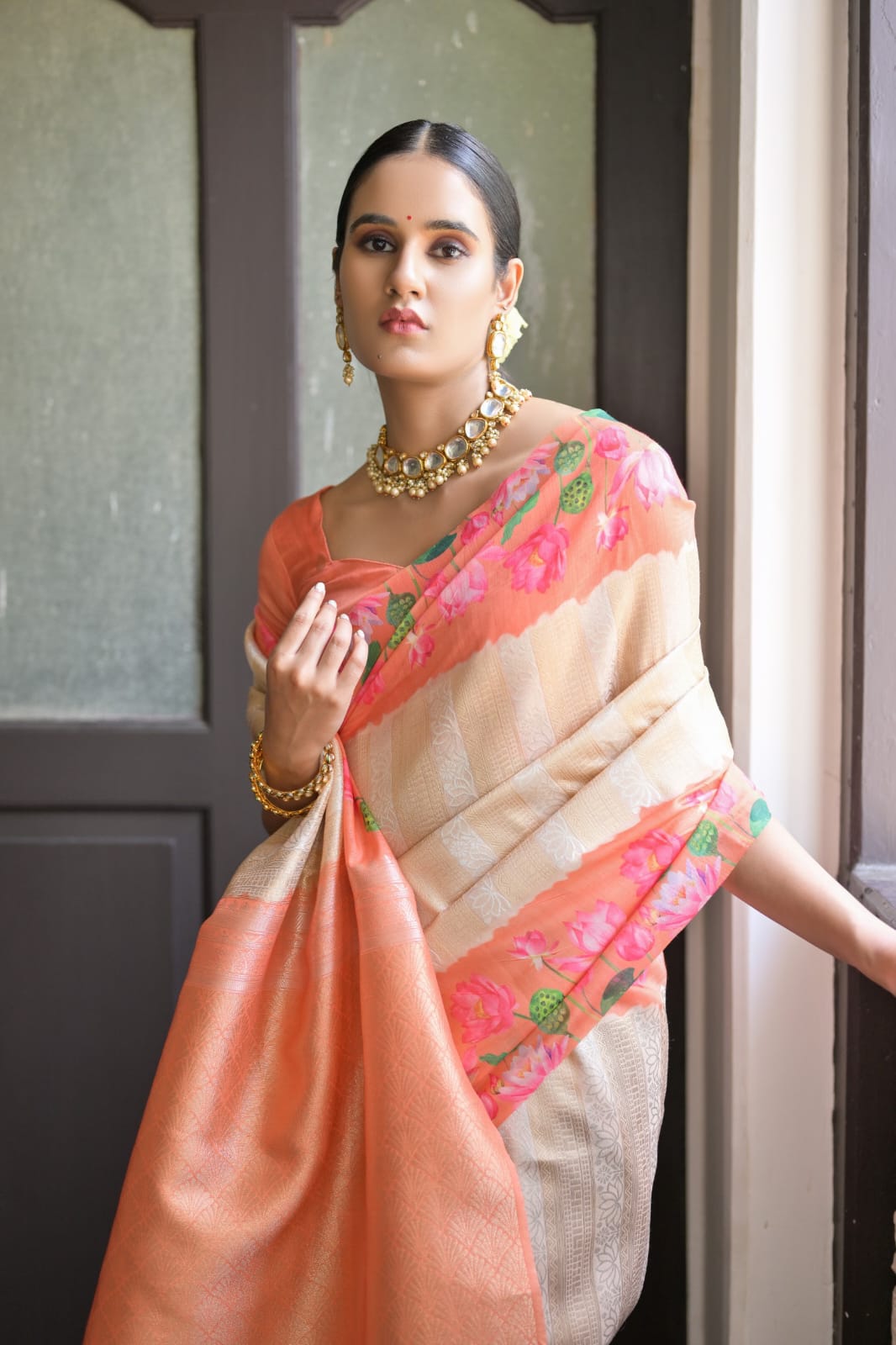 Beautiful Indian woman in a soft peach zari work silk saree, adorned with traditional jewelry, standing by a window.