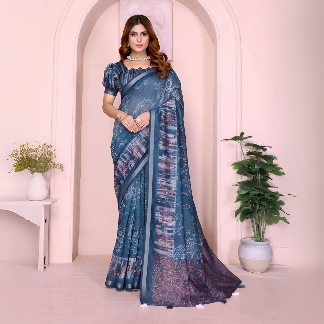 Woman wearing a steel blue printed saree with intricate abstract and paisley designs, paired with a matching short-sleeve blouse and accessorized with a silver choker necklace, bangles, and rings. She poses gracefully in a modern studio featuring a pastel arched alcove, a white side table with a ceramic vase of green foliage, and a potted plant against a soft blush background.