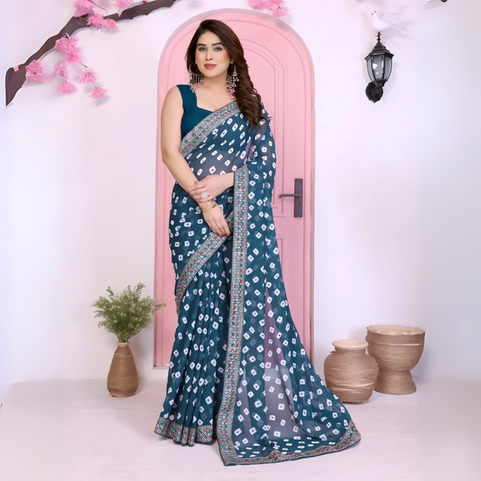 Woman wearing a teal bandhani saree with traditional white tie-dye patterns and a decorative embroidered border, paired with a contrasting blue blouse. She stands gracefully in front of a pastel pink arched doorway, accessorized with statement earrings and bangles. The contemporary studio setting features earthy clay pots, a small green foliage vase, and decorative pink branches, creating a modern minimalist backdrop.