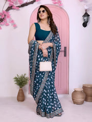 Woman wearing a teal bandhani saree with traditional white tie-dye patterns and a decorative embroidered border, paired with a contrasting blue blouse. She stands gracefully in front of a pastel pink arched doorway, accessorized with statement earrings and bangles. The contemporary studio setting features earthy clay pots, a small green foliage vase, and decorative pink branches, creating a modern minimalist backdrop.