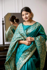 Woman wearing a teal silk saree with intricate brocade patterns and a matching blouse, standing gracefully beside a vintage black dresser with a mirror in a softly lit studio featuring white paneled walls and elegant decor.