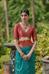 Woman wearing a teal and coral silk saree with gold zari motifs and a contrasting coral border, posing outdoors on a vintage stone veranda with lush green trees and garden plants in the background.
