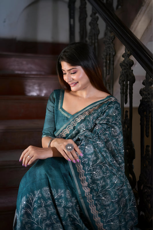 Woman wearing a teal floral printed saree with a matching blouse, standing beside a vintage black table in a heritage-style bedroom with a green paneled door and antique wooden furniture.