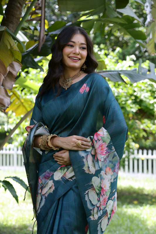 Woman wearing a teal saree with large handpainted lotus and leaf floral motifs, accessorized with traditional gold jewelry, standing on a lush green lawn surrounded by tropical plants and a white picket fence in the background.