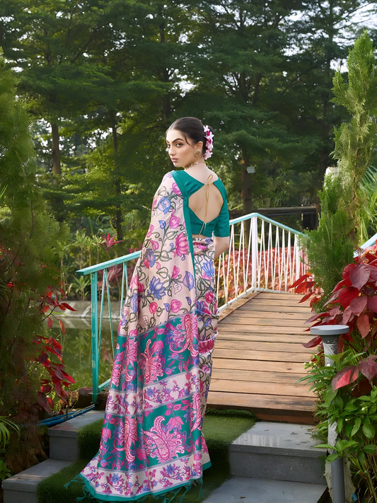 Woman wearing a teal and pink saree with vibrant Madhubani-inspired floral print border and pallu, paired with a teal blouse and traditional jewelry, gracefully standing on stone steps beside a wooden bridge surrounded by lush greenery and colorful garden plants.