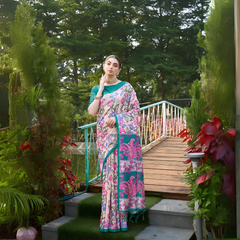 Woman wearing a teal and pink saree with vibrant Madhubani-inspired floral print border and pallu, paired with a teal blouse and traditional jewelry, gracefully standing on stone steps beside a wooden bridge surrounded by lush greenery and colorful garden plants.