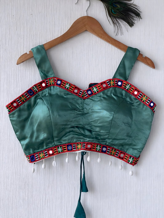 Teal green ethnic blouse featuring vibrant mirror work and a multicolored embroidered geometric border in red, green, blue, and white, adorned with white pearl drop tassels and a tie-up waist detail. Displayed on a wooden hanger against a textured off-white wall with peacock feather, creating a festive and traditional look.