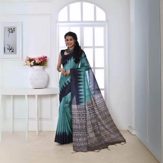 Woman wearing a teal and navy blue Ikat printed saree with geometric patterns on the pallu and border, paired with a navy blouse and statement necklace, standing in a modern white interior with large arched windows and a white console table decorated with a vase of pink flowers.