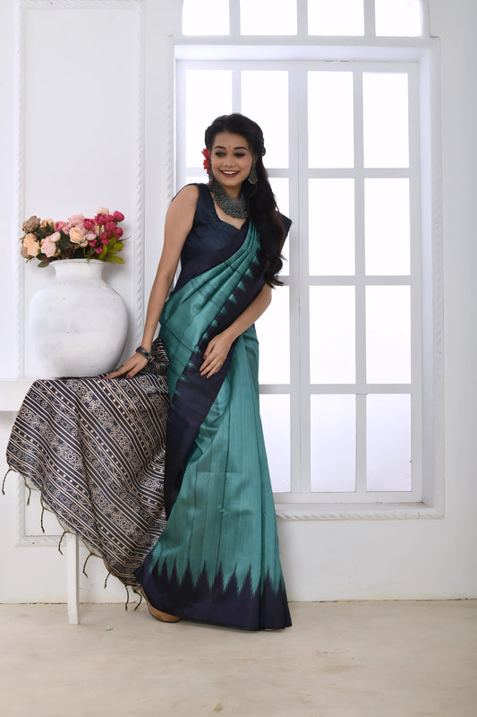 Woman wearing a teal and navy blue Ikat printed saree with geometric patterns on the pallu and border, paired with a navy blouse and statement necklace, standing in a modern white interior with large arched windows and a white console table decorated with a vase of pink flowers.