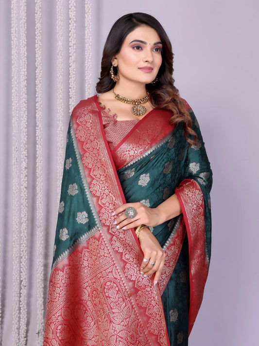 Woman draped in an elegant teal Banarasi silk saree adorned with intricate silver woven motifs and a rich red zari border, paired with a matching red blouse and classic gold jewelry. She poses gracefully in a contemporary studio decorated with woven baskets filled with rose petals, sheer white curtains, and a minimalist neutral backdrop.