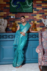 Woman wearing an elegant teal silk saree with intricate gold woven border and floral motifs, paired with traditional gold jewelry, gracefully posing in a vintage-inspired blue interior with artistic wall decor, a sculpted statue, and patterned armchair.