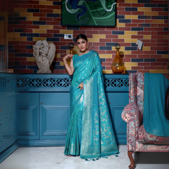 Woman wearing an elegant teal silk saree with intricate gold woven border and floral motifs, paired with traditional gold jewelry, gracefully posing in a vintage-inspired blue interior with artistic wall decor, a sculpted statue, and patterned armchair.