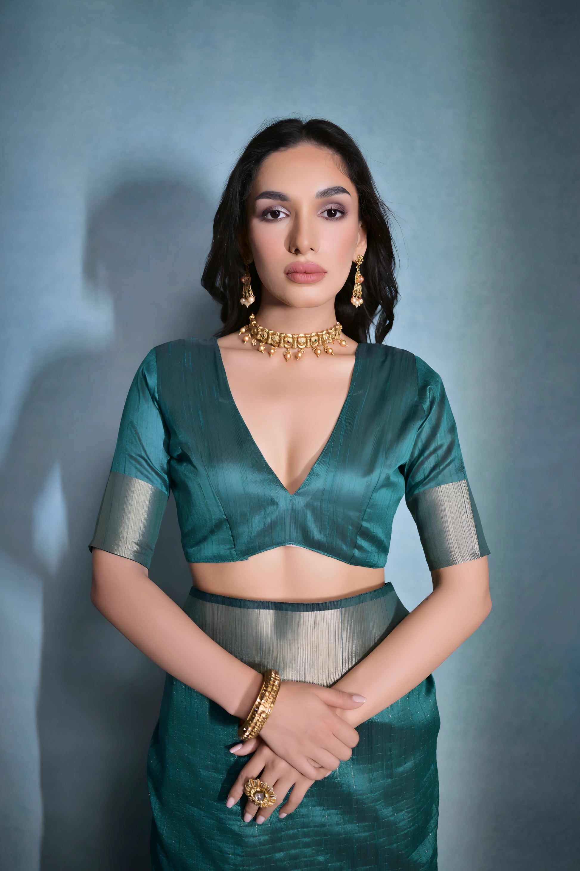  Woman wearing a teal silk saree with a gold border and traditional jewelry, standing against a blue background