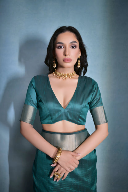  Woman wearing a teal silk saree with a gold border and traditional jewelry, standing against a blue background