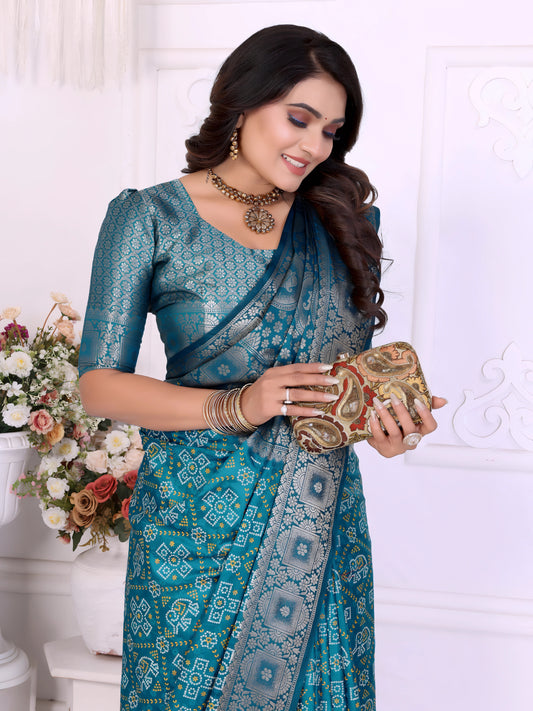 Woman wearing a teal blue silk saree with intricate silver zari border and traditional floral motifs, paired with a matching blouse and statement jewelry. She poses gracefully in a classic white room decorated with paneled walls and white vases filled with pastel floral arrangements.