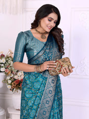 Woman wearing a teal blue silk saree with intricate silver zari border and traditional floral motifs, paired with a matching blouse and statement jewelry. She poses gracefully in a classic white room decorated with paneled walls and white vases filled with pastel floral arrangements.