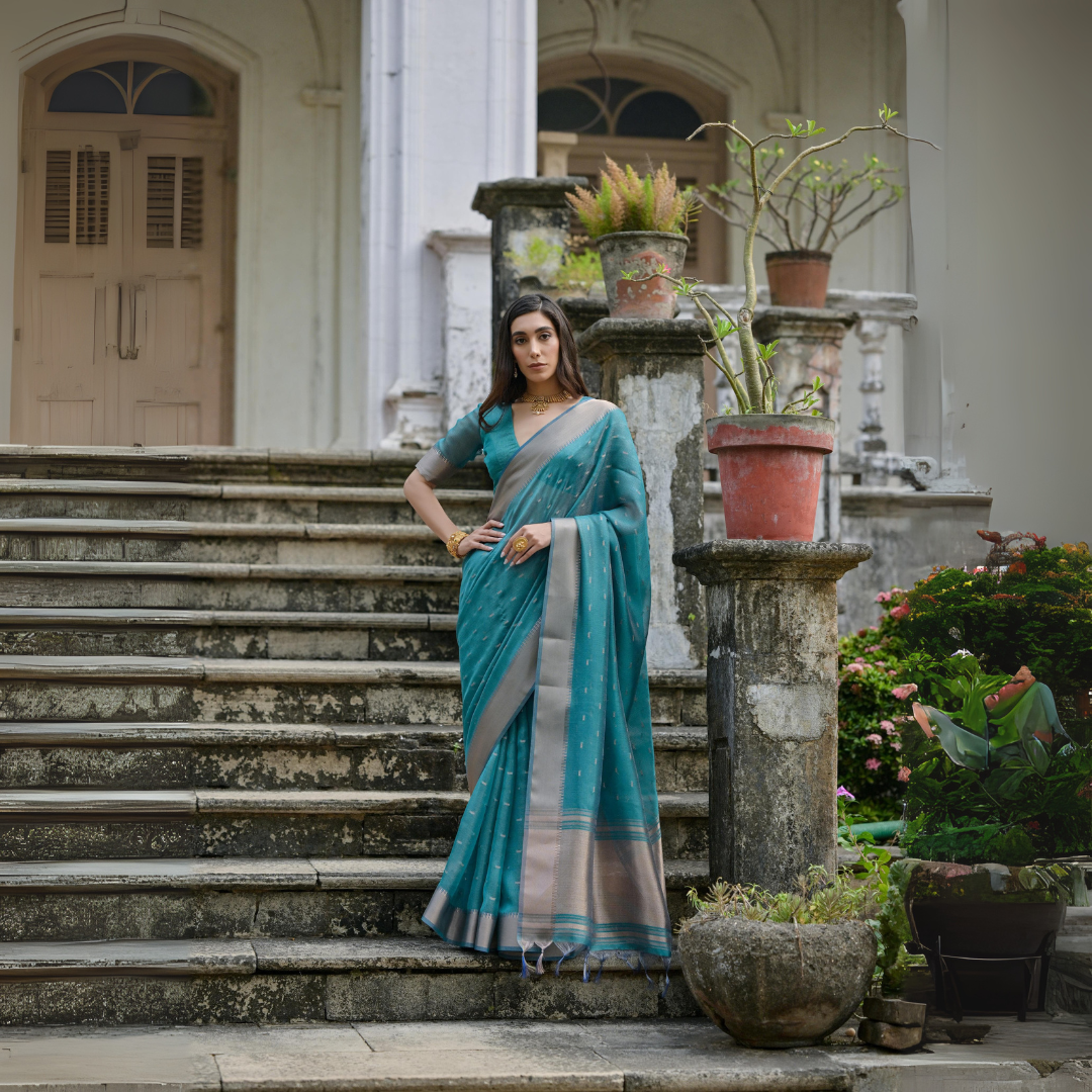 Woman wearing a teal silk saree with a silver border and subtle woven motifs, paired with a matching blouse and gold jewelry, standing on the steps of a heritage mansion surrounded by potted plants and lush greenery.