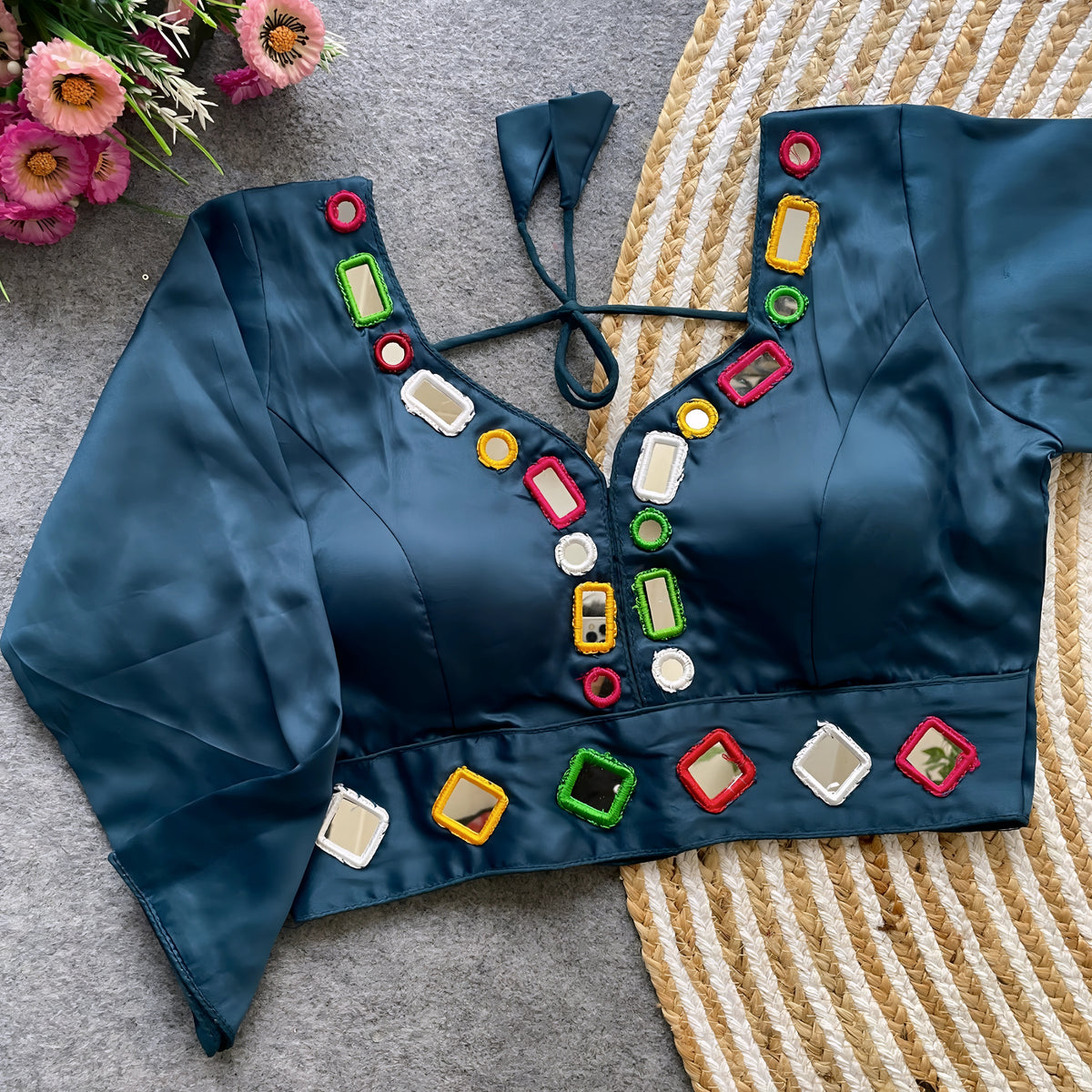 Teal blouse adorned with square and round mirror work framed by multicolored embroidered borders, stylish back tie-up closure, and three-quarter sleeves. Displayed flat on a textured grey surface with a woven mat and fresh flowers, highlighting its festive and contemporary ethnic design.