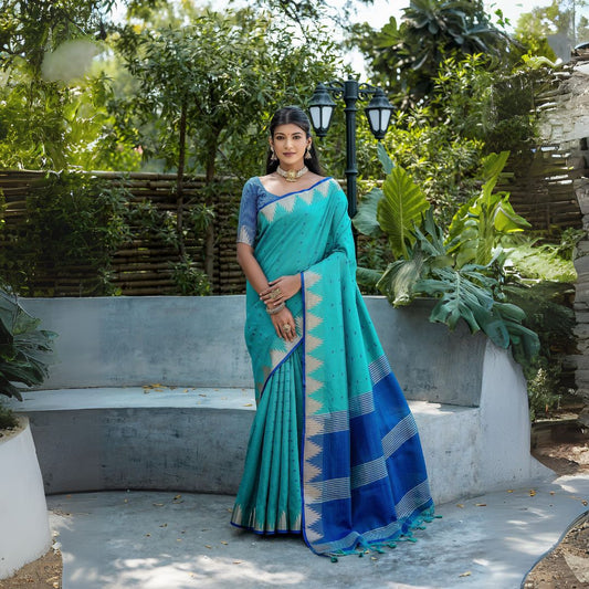 A woman models a striking Teal Blue Tussar Silk Saree with small woven motifs. The saree has a beige/silver Zari temple border and a pallu featuring wide bands of deep royal blue and Zari stripes. She wears a printed blue blouse and heavy gold Kundan jewelry.
