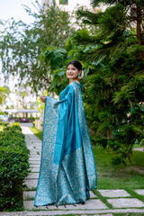 Woman wearing a turquoise blue silk saree with intricate silver brocade patterns and a matching blouse, accessorized with statement earrings and bangles, standing gracefully on a stone pathway in a lush green garden with manicured hedges and tropical trees in the background.