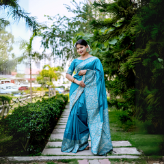 Woman wearing a turquoise blue silk saree with intricate silver brocade patterns and a matching blouse, accessorized with statement earrings and bangles, standing gracefully on a stone pathway in a lush green garden with manicured hedges and tropical trees in the background.