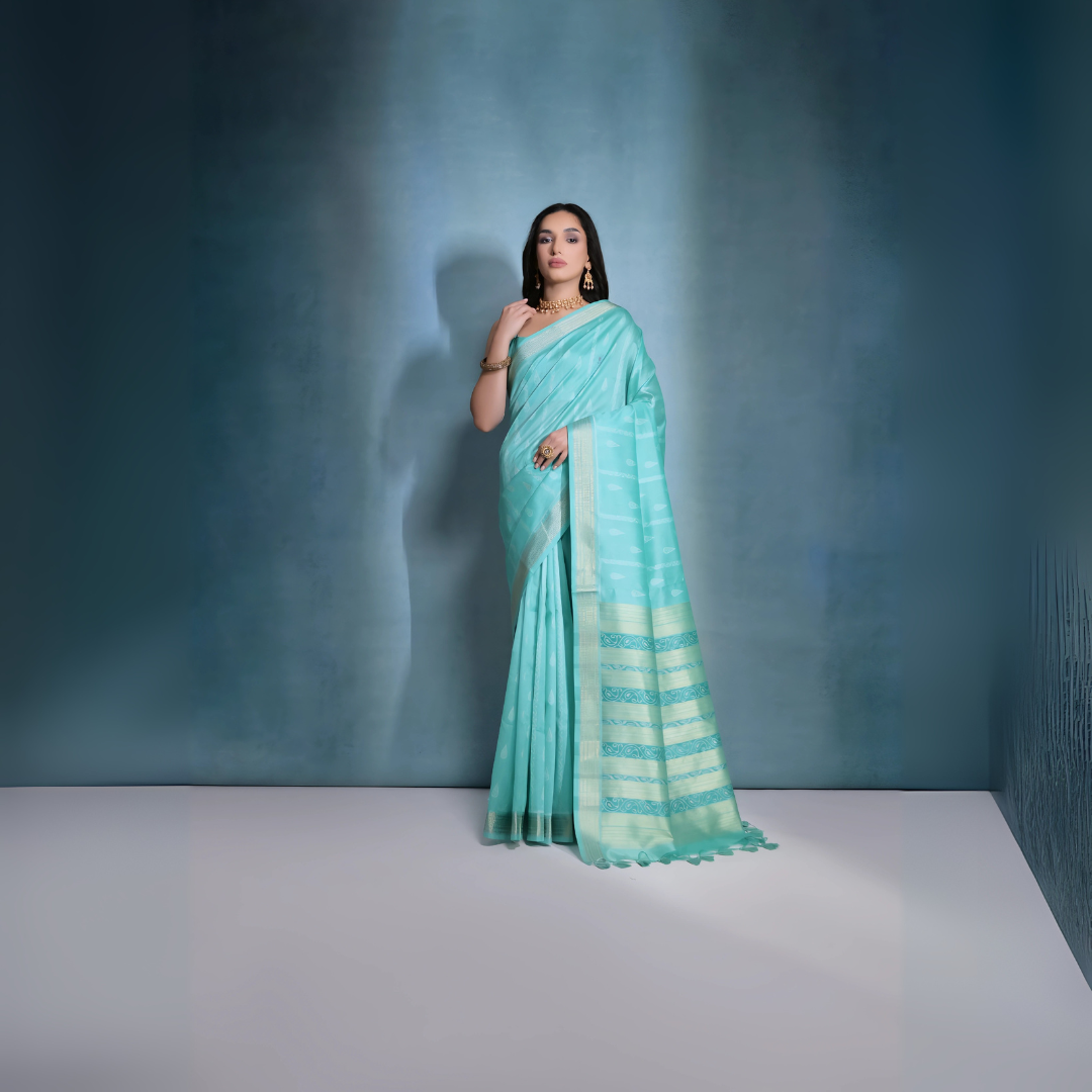 Woman wearing a turquoise silk saree with gold zari border and intricate patterns, paired with a matching blouse and traditional gold jewelry including a choker necklace and earrings, posing elegantly in a studio setting with a soft blue-grey gradient background.