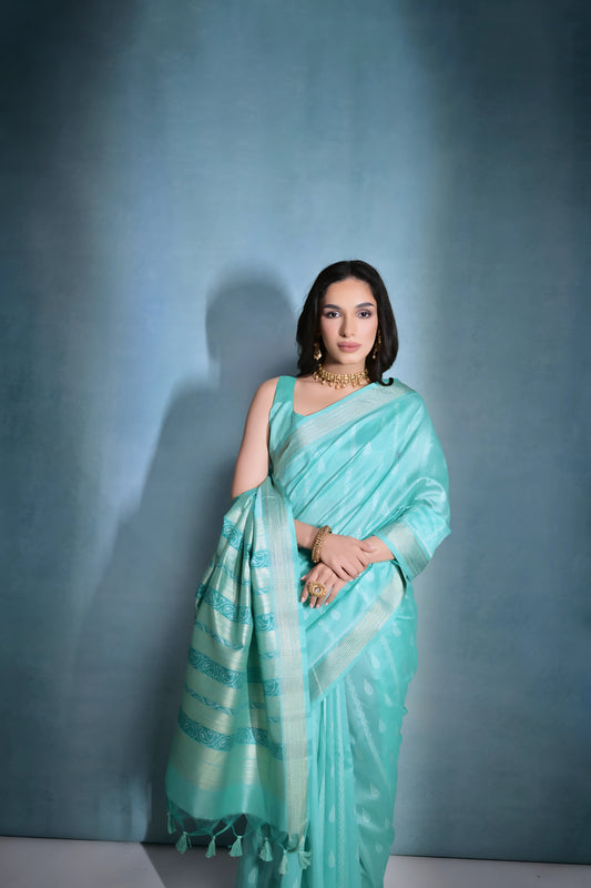 Woman wearing a turquoise silk saree with gold zari border and intricate patterns, paired with a matching blouse and traditional gold jewelry including a choker necklace and earrings, posing elegantly in a studio setting with a soft blue-grey gradient background.