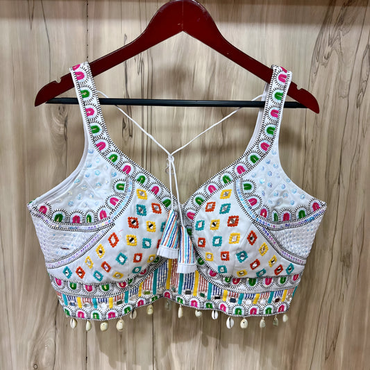 Traditional white Gujarati blouse featuring vibrant multicolor geometric embroidery in green, pink, yellow, orange, and blue, accented with intricate mirror work and white cowrie shell trim along the hem. The design includes wide embroidered straps, structured cups, tasseled tie-back detail, and is displayed on a wooden hanger against a light wood panel background.